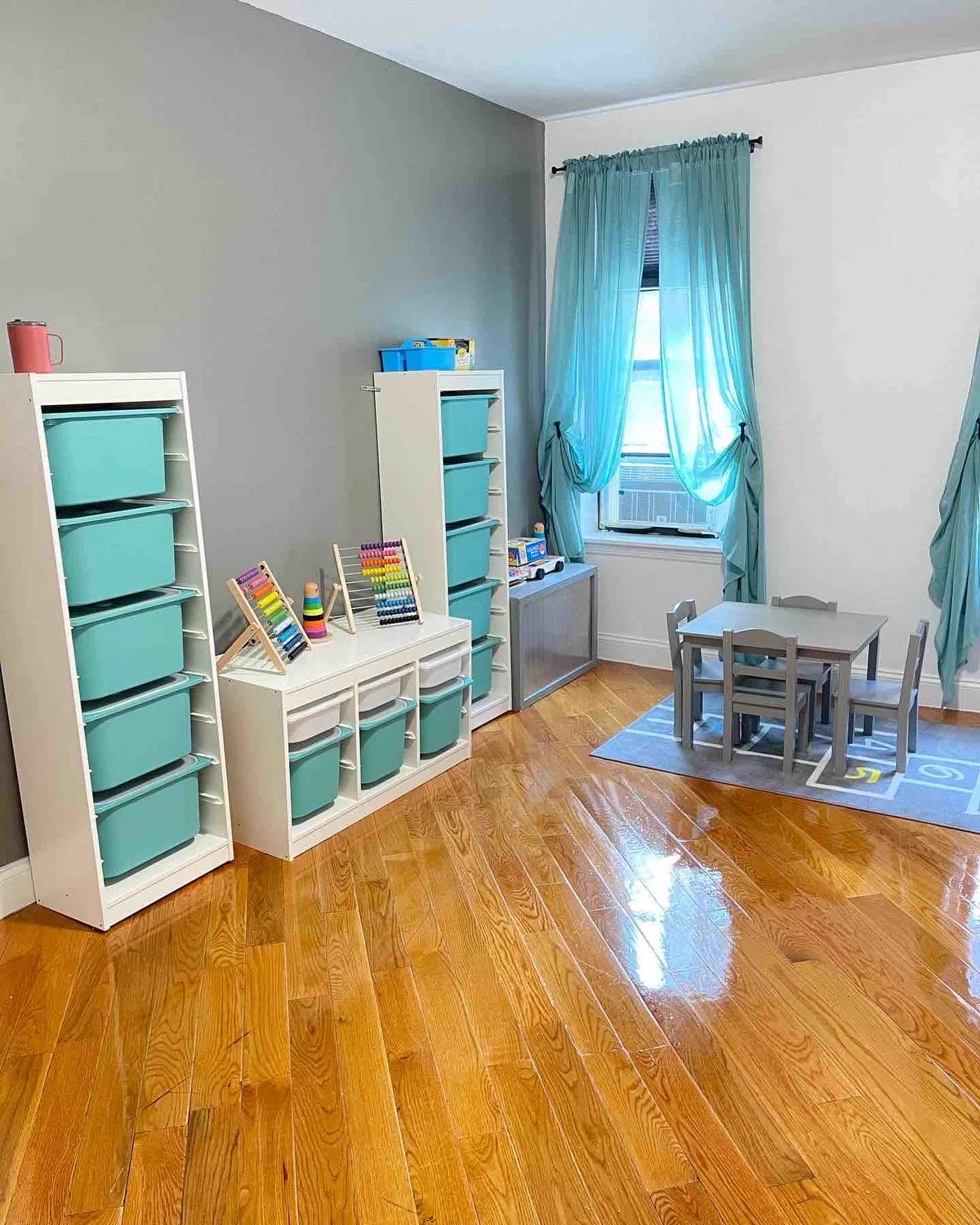 Área de juego de Creative Minds: estantes turquesa a la altura de los niños, un ábaco de colores, mesita y cortinas turquesa junto a la ventana