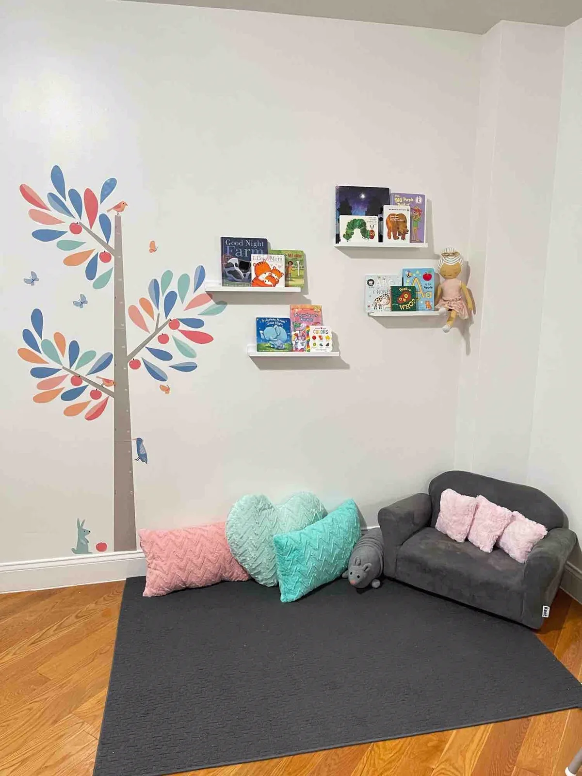 Rincón de lectura de Creative Minds: libros en repisas, un árbol pintado en la pared y cojines en forma de corazón