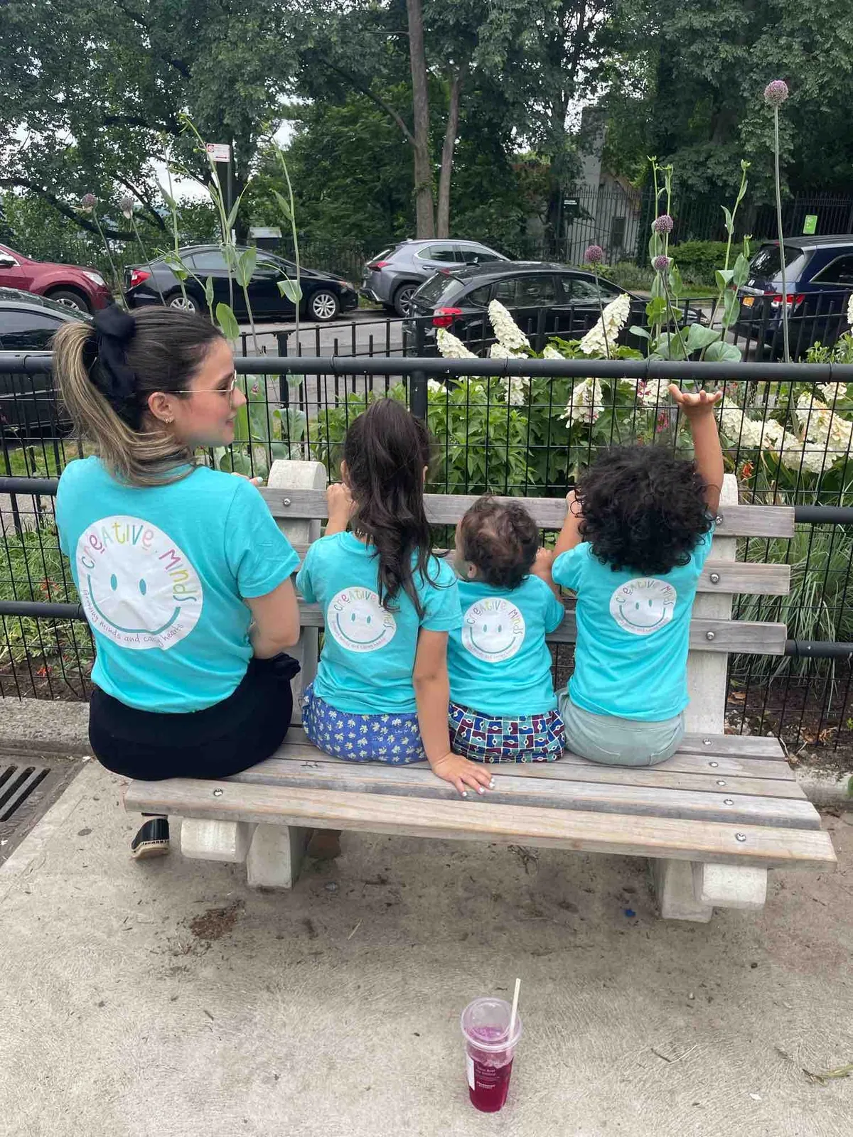 Una maestra y tres niños de Creative Minds, de espaldas y con camisetas turquesa del daycare, sentados en un banco frente a un jardín